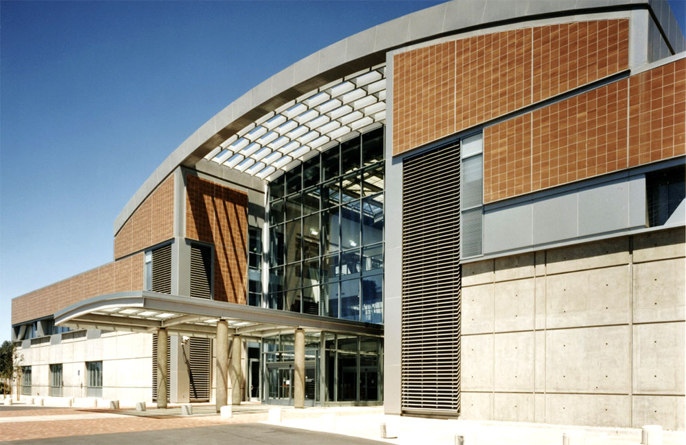 Image 4. Front view of building entrance showing concrete walls on first floor and terra cotta-colored tiles on the upper levels. A steel and glass canopy extends from the main doors towards the parking lot.
