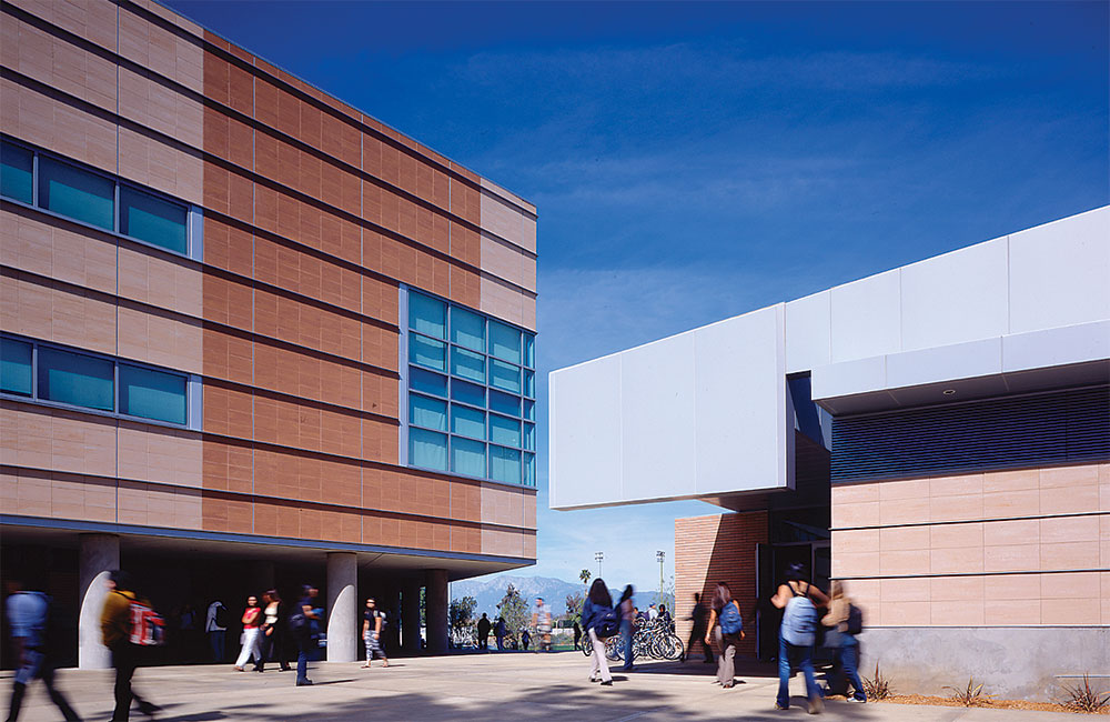 Image 4. A courtyard framed by a 3-story sandstone-colored building on the left and a 1-story sandstone-colored building with angular light grey metal roof on the right.