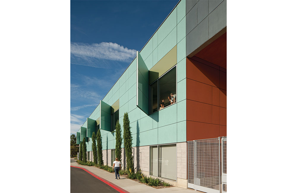 Image 2. Side view of 2-story building showing turquoise-colored panels on upper level and concrete colored tiles on the first floor. Vertical sunshade panels pivot outwardly from the second-floor windows. 