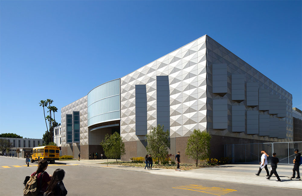 Image 1. Corner exterior view of 3-story modern building clad with metal panels on the second and third levels.  Vertical angled window slots punctuate the facade in the upper floors providing sunlight and views to the interior rooms.